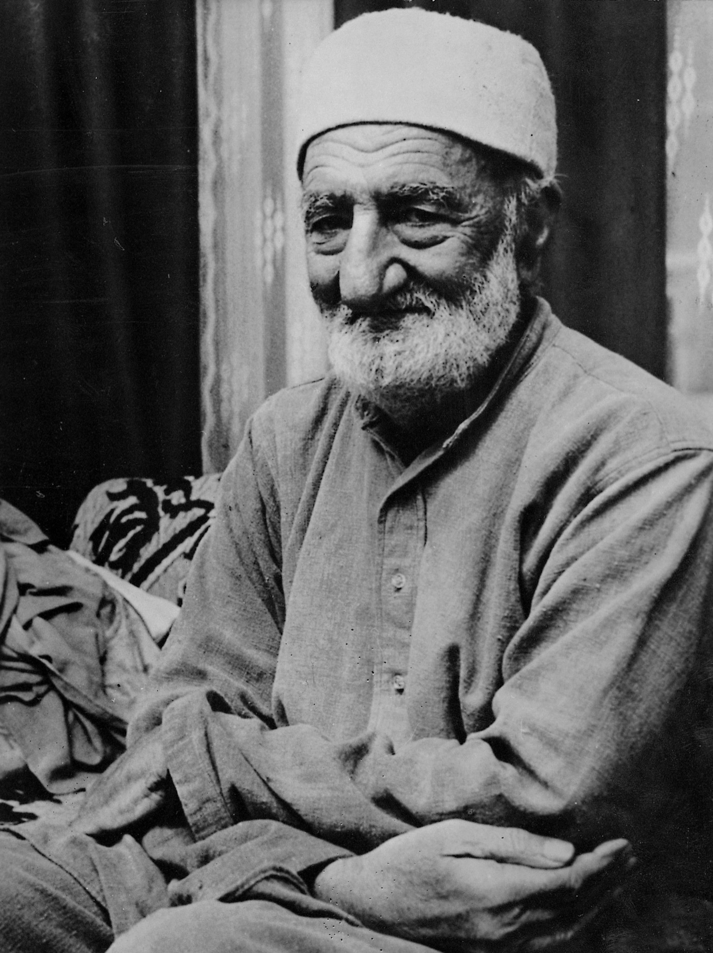 Khan Abdul Ghaffar Khan in later life, seated cross-legged in a plain kurta and white cap, hands resting in his lap.