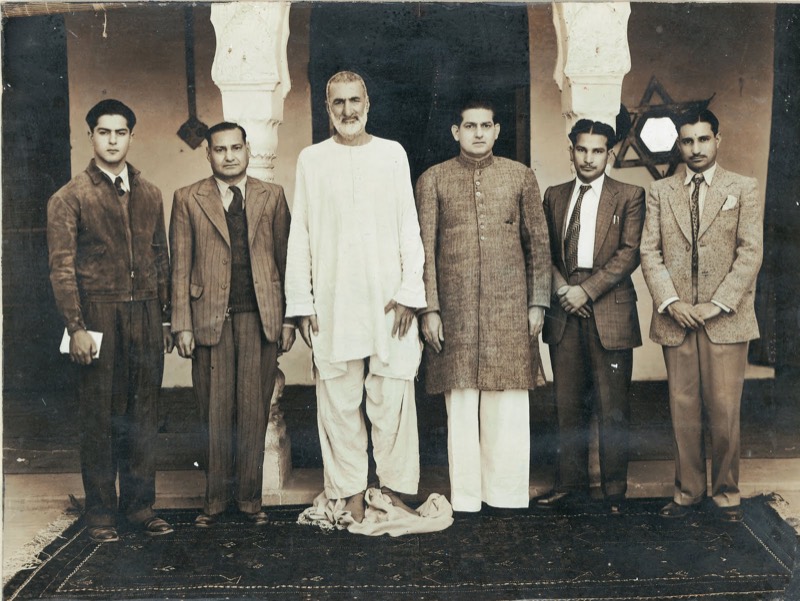 Sepia group photograph: Ghaffar Khan at the centre, tall and barefoot in plain white homespun, flanked by six men in suits and a sherwani, on a patterned carpet before an arched colonnade.