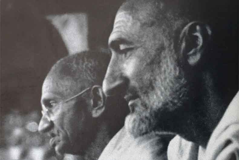 Black and white photograph in profile: Khan Abdul Ghaffar Khan in the foreground on the right, bearded, looking ahead; Mahatma Gandhi behind him on the left in spectacles, also in profile, at a public gathering.