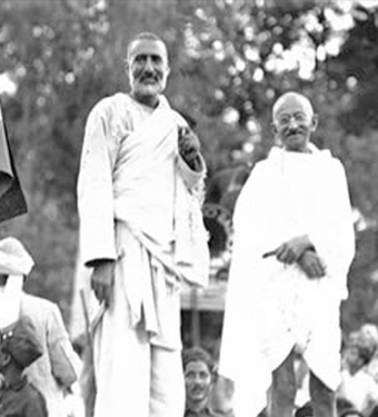 Black and white photograph of a public gathering: Ghaffar Khan standing at his full height in white, with Mahatma Gandhi beside him in a shawl, a crowd around them.